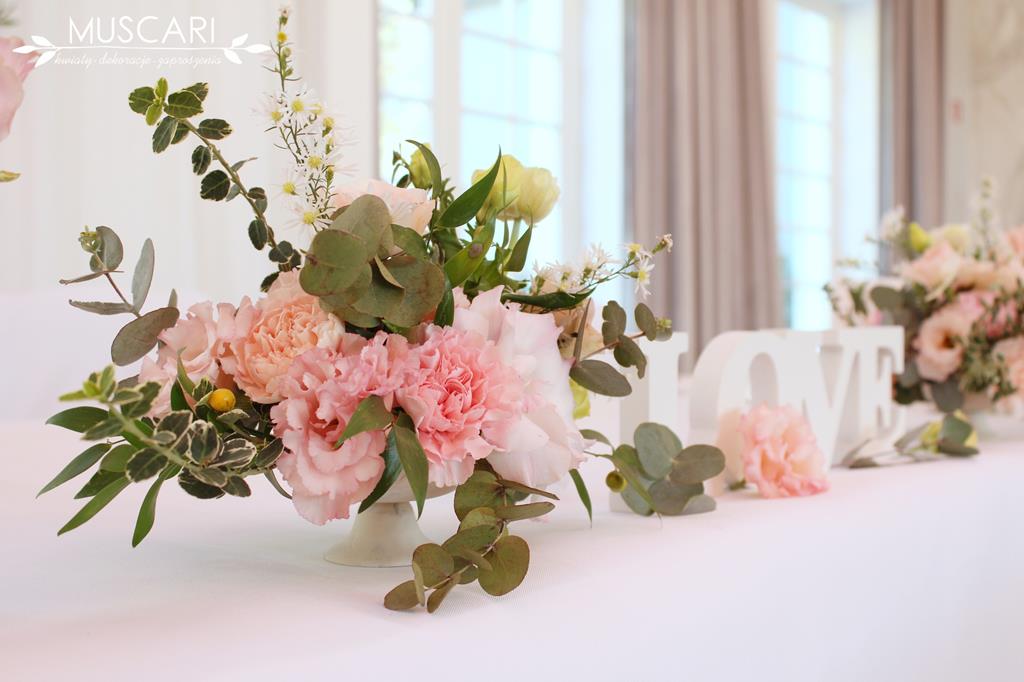 pastelowo różowa kompozycja z kwiatów ozobiona zielenią na stole pary młodej